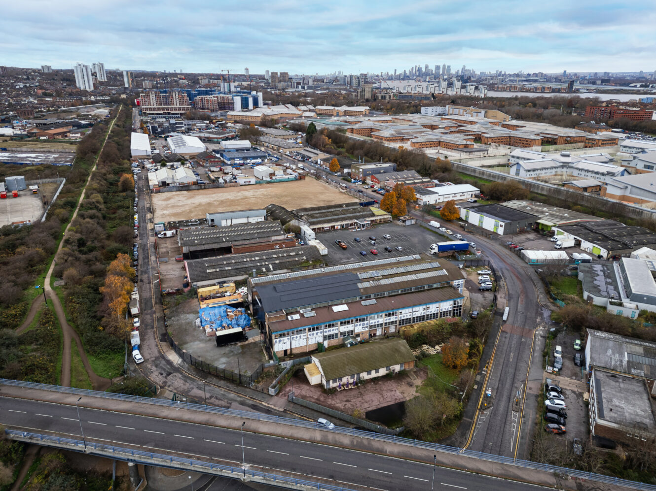 Aerial view of an industrial area with warehouses, roads, parked vehicles, and city buildings in the distant background under a cloudy sky.