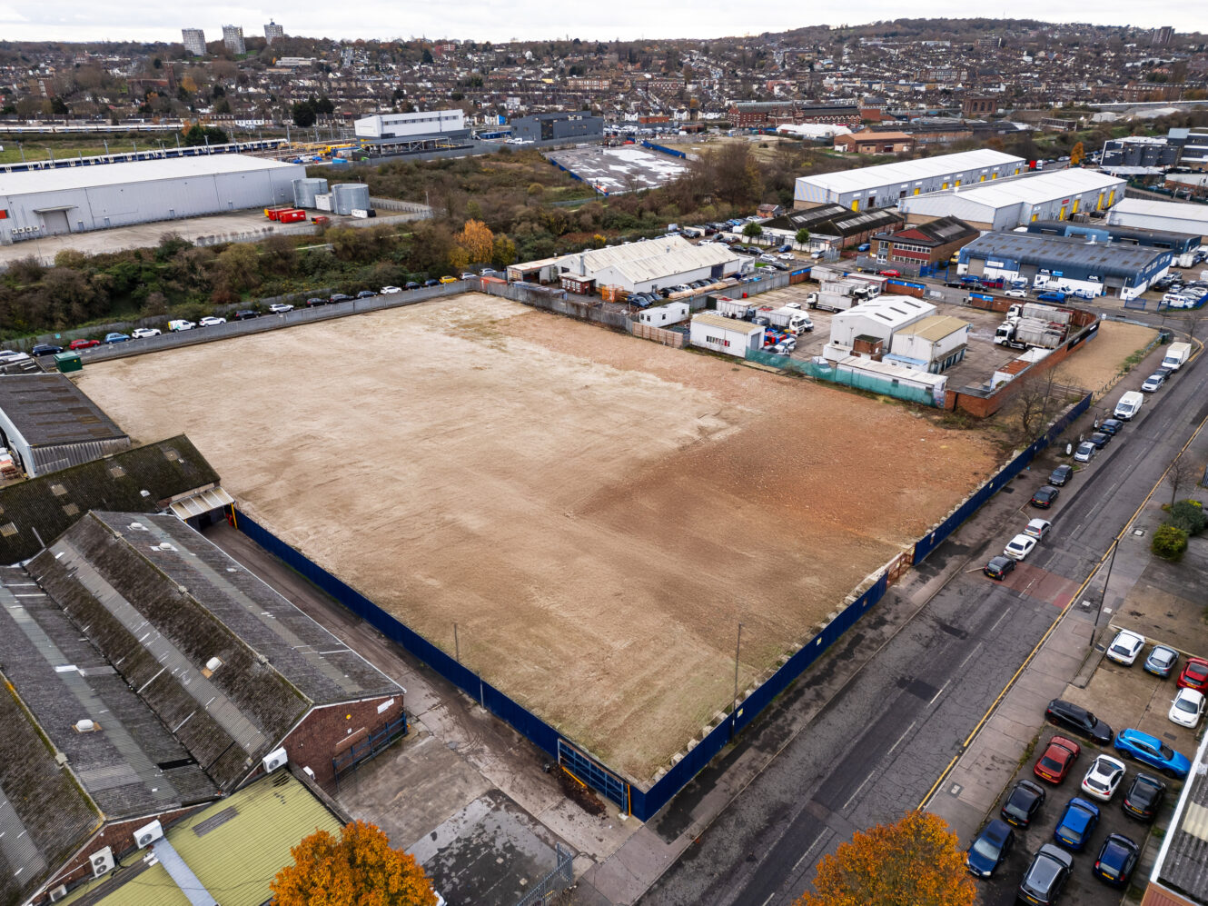 A large, fenced, cleared plot of land is surrounded by industrial buildings, warehouses, and a few parked vehicles in an urban area.