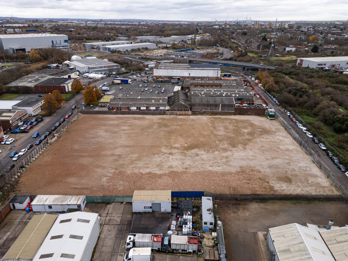 A large, empty, rectangular lot bordered by industrial buildings and warehouses, with roads and vehicles surrounding the area under a cloudy sky.