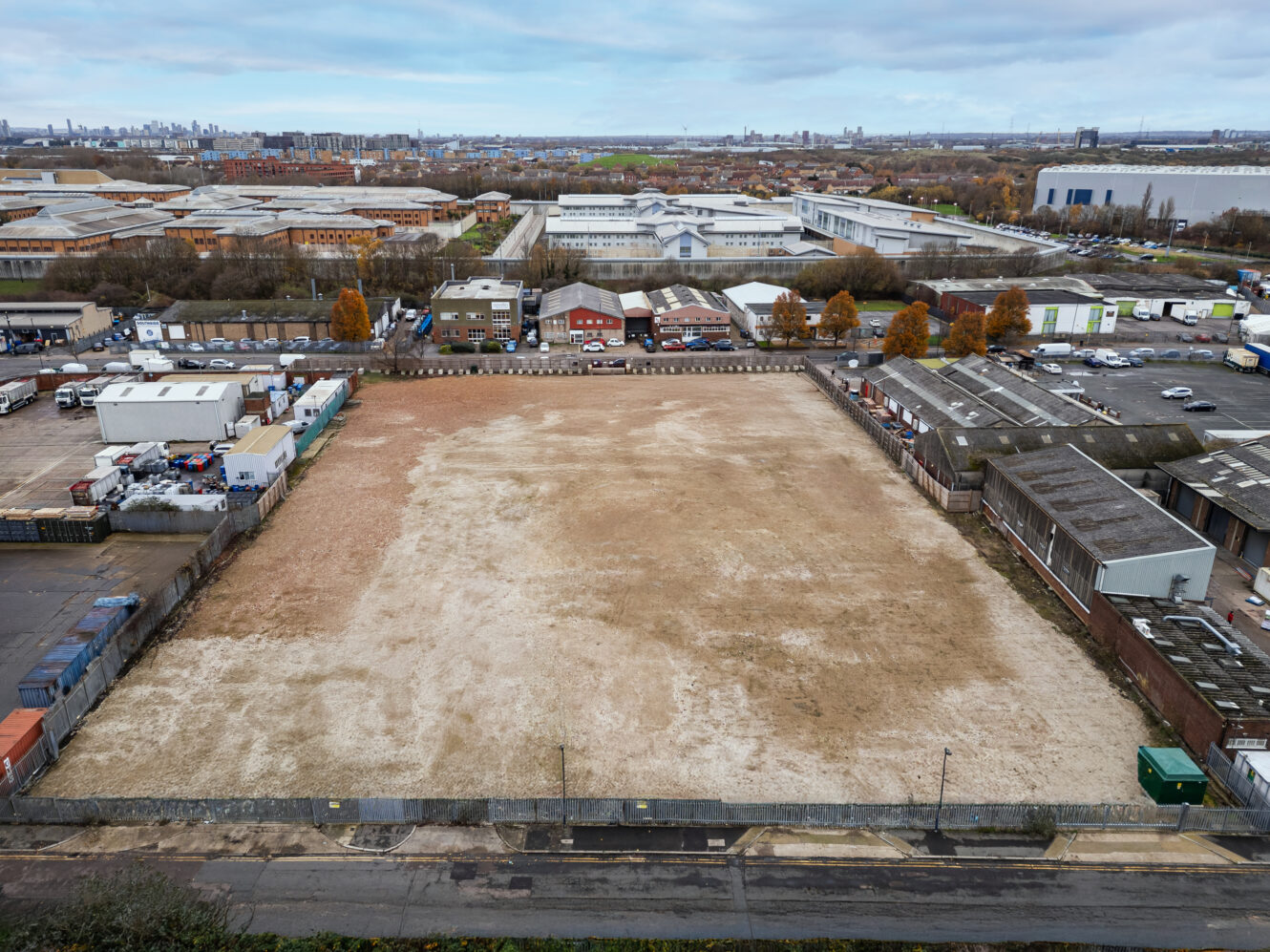 A large vacant dirt lot surrounded by industrial warehouses and buildings, with a cityscape visible in the distant background under a cloudy sky.