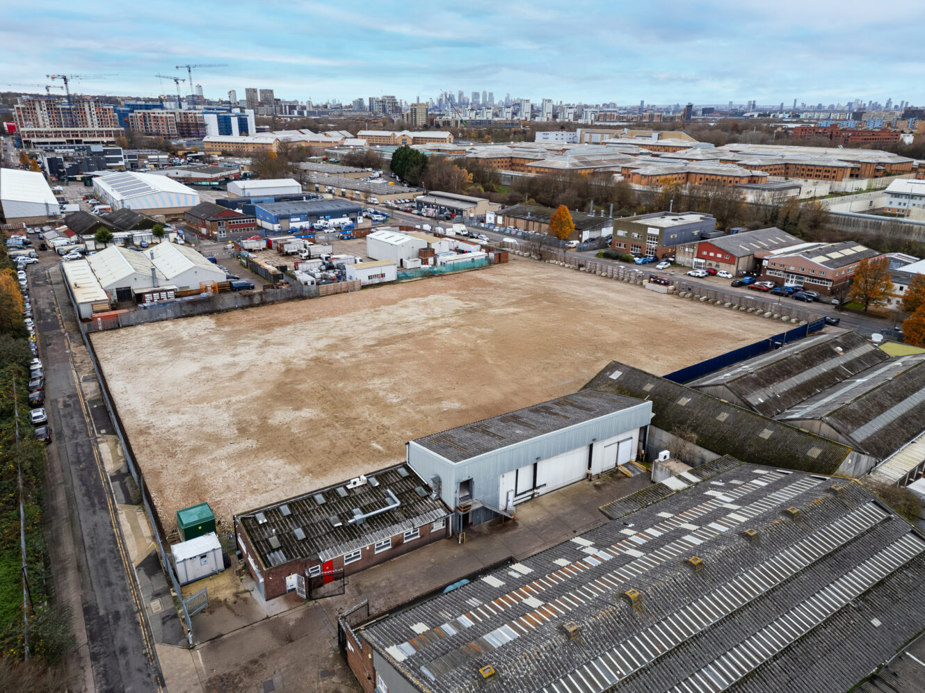 Aerial view of an empty, fenced industrial plot surrounded by warehouses and industrial buildings in an urban area.