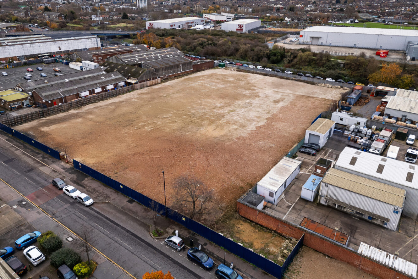 A large, empty dirt lot is surrounded by industrial buildings and a blue fence, with parked cars along the adjacent street.