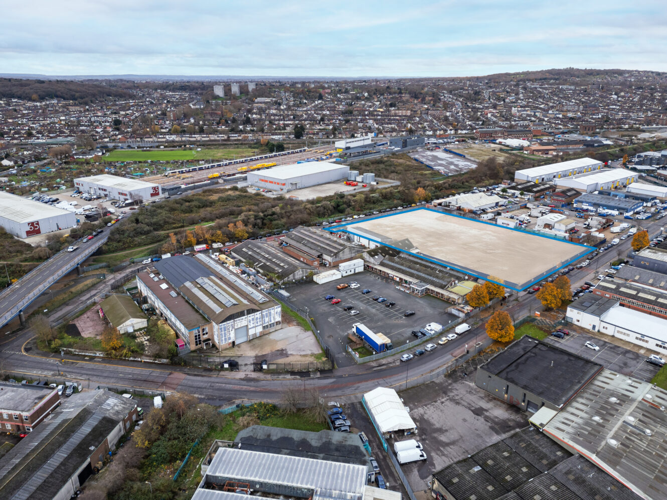 Aerial view of an industrial estate with warehouses, parking lots, roads, and some trees; residential areas and hills visible in the background under a cloudy sky.