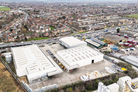 Aerial view of an industrial complex with large warehouses, nearby roads, parked vehicles, and a residential area in the background.