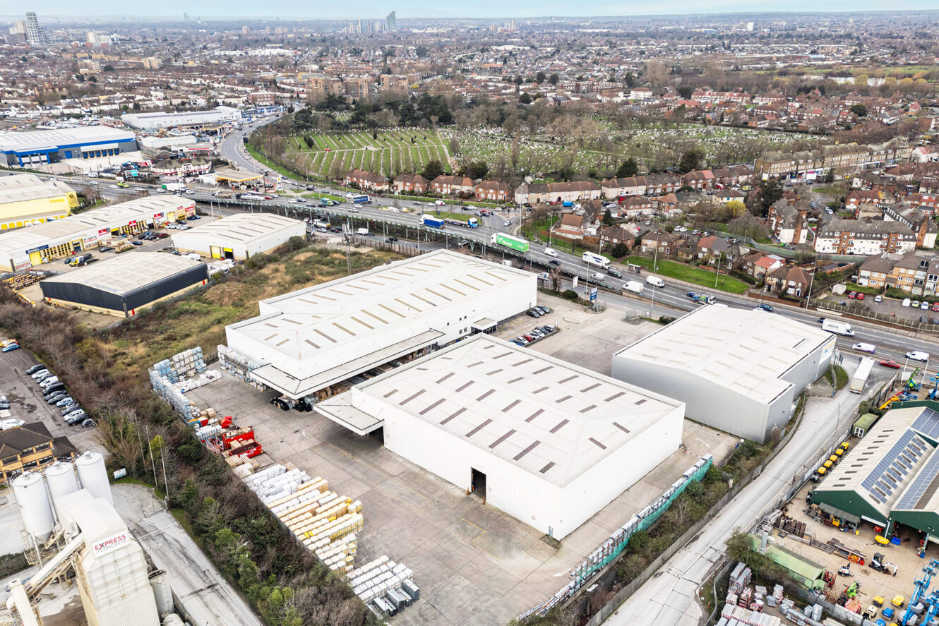 Aerial view of an industrial complex with large warehouse buildings, outdoor storage, surrounding roads, and residential area in the background.