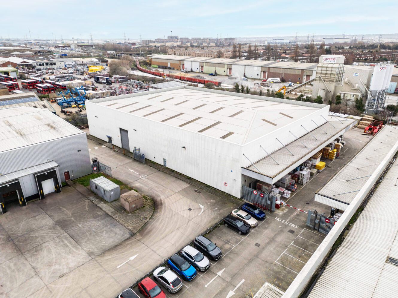 Aerial view of a large warehouse with loading bays, parked cars in a lot, and surrounding industrial buildings under a cloudy sky.