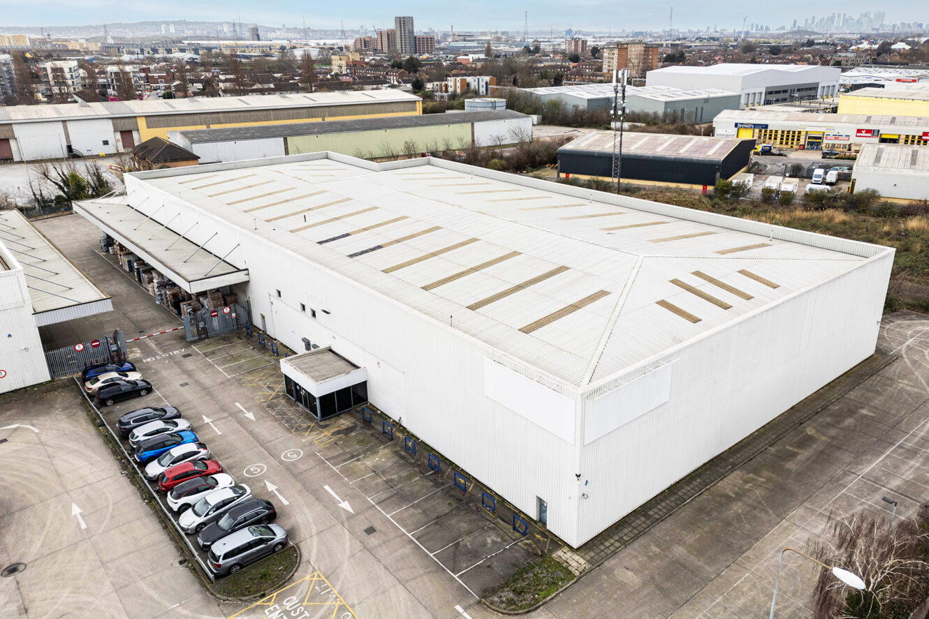 Aerial view of a large white industrial warehouse with parked cars outside, located in an urban commercial area with other warehouses and buildings nearby.