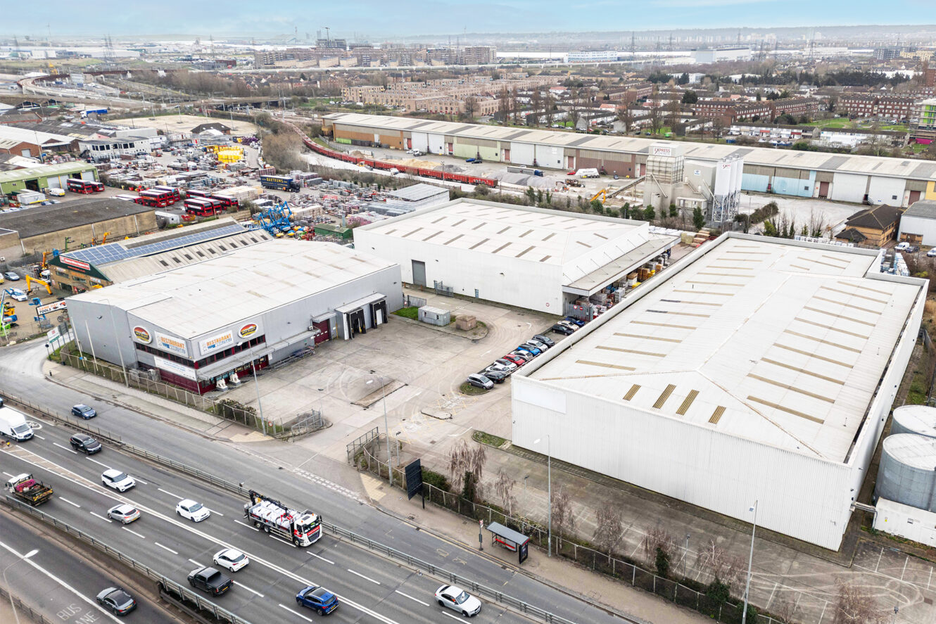 Aerial view of an industrial estate with warehouses, parked vehicles, and a busy road in the foreground; cityscape visible in the background.