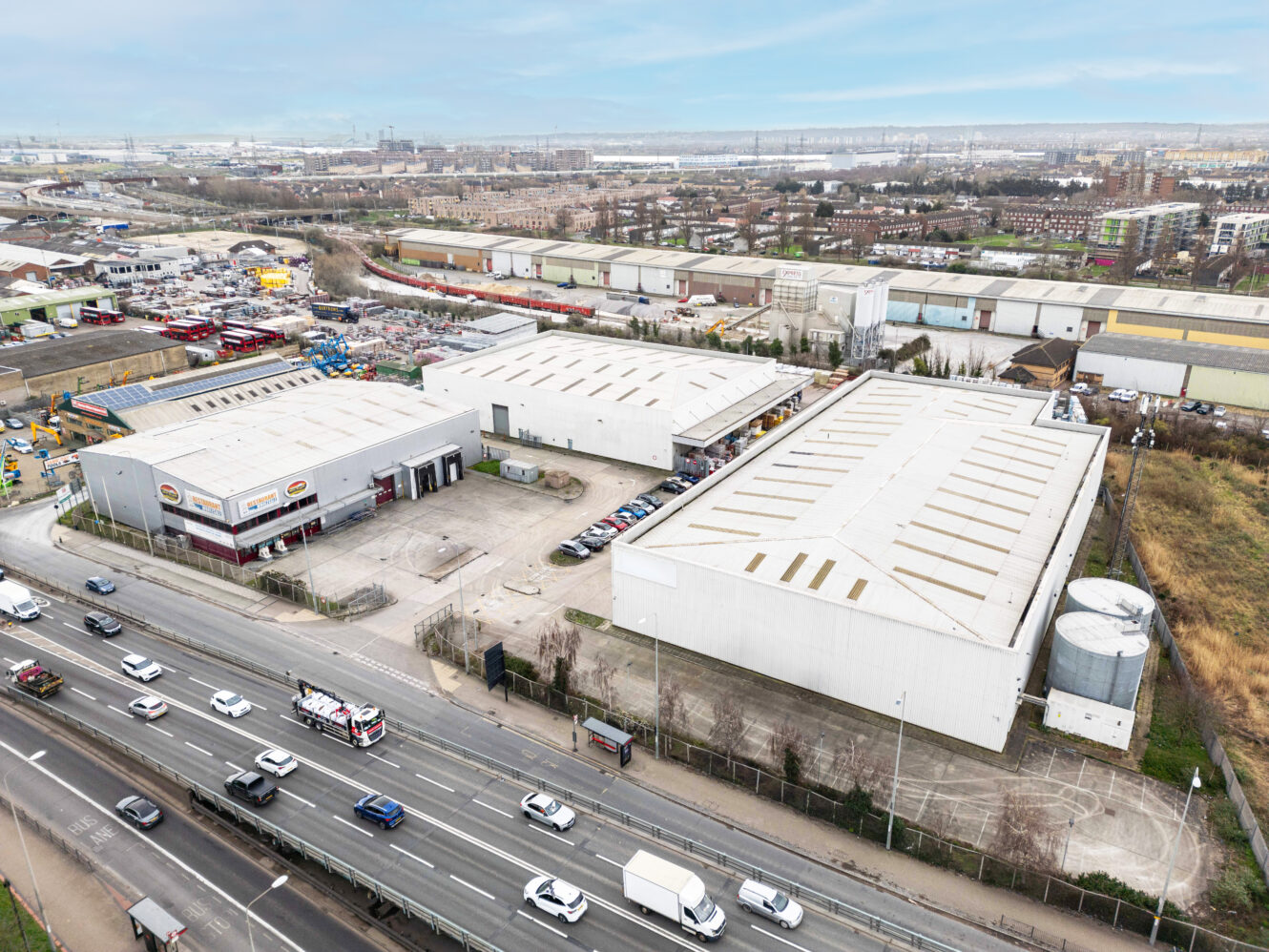 Aerial view of industrial warehouses and parking lots next to a busy multi-lane road, with cityscape and factories in the background.