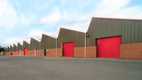 A row of industrial warehouse units with red roller doors, brick lower walls, and green upper façades under a partly cloudy sky.