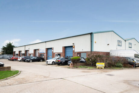 A row of industrial warehouse units with blue roller shutter doors, several parked cars, and a small landscaped area under a clear sky.