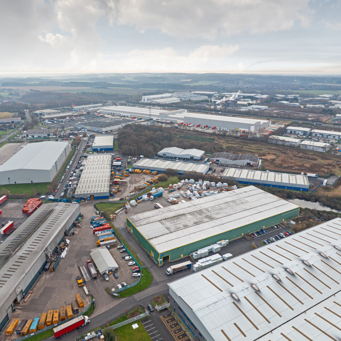 Aerial view of an industrial estate with multiple large warehouses, parked vehicles, and surrounding roads, set in a semi-rural landscape under a cloudy sky.