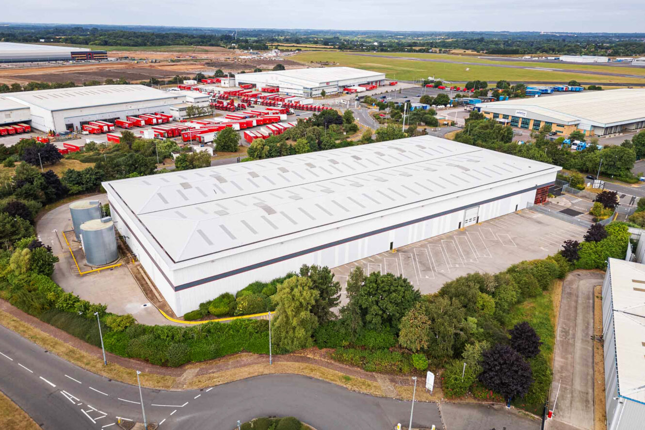 Aerial view of a large industrial warehouse with a white roof, surrounded by trees and parking space, located in an industrial park with other warehouses and red vehicles.