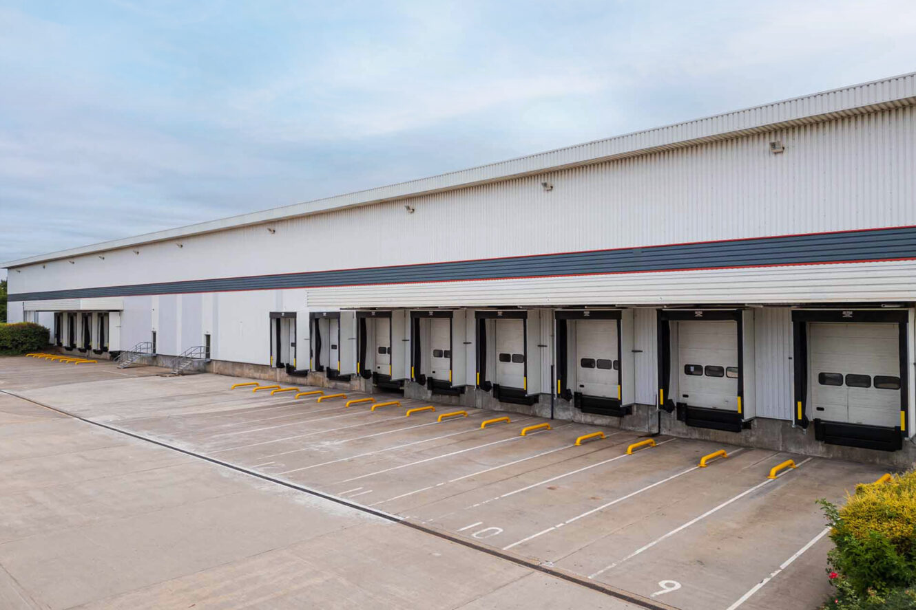 A large warehouse with multiple closed loading dock doors lined up along its side and empty numbered parking bays in front.