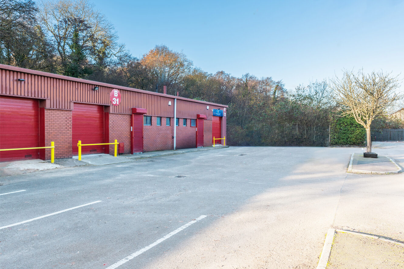 Industrial building with red doors and windows, yellow bollards in front, and an empty parking lot bordered by trees and a single leafless tree.