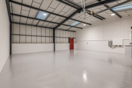 Empty industrial warehouse space with clean gray floor, white walls, overhead lights, metal ceiling, skylights, and a red exit door at the back.