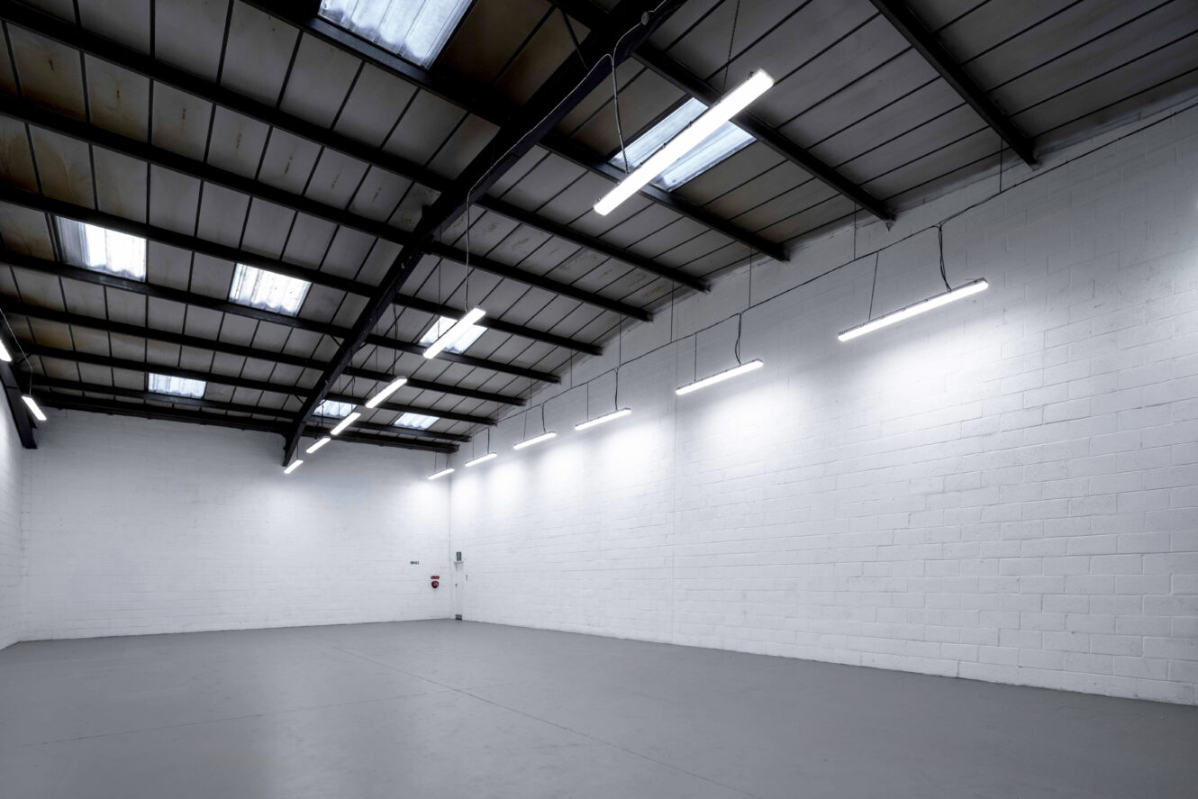 Empty industrial warehouse space with white brick walls, gray concrete floor, and fluorescent ceiling lights. Skylights allow natural light in from the roof.