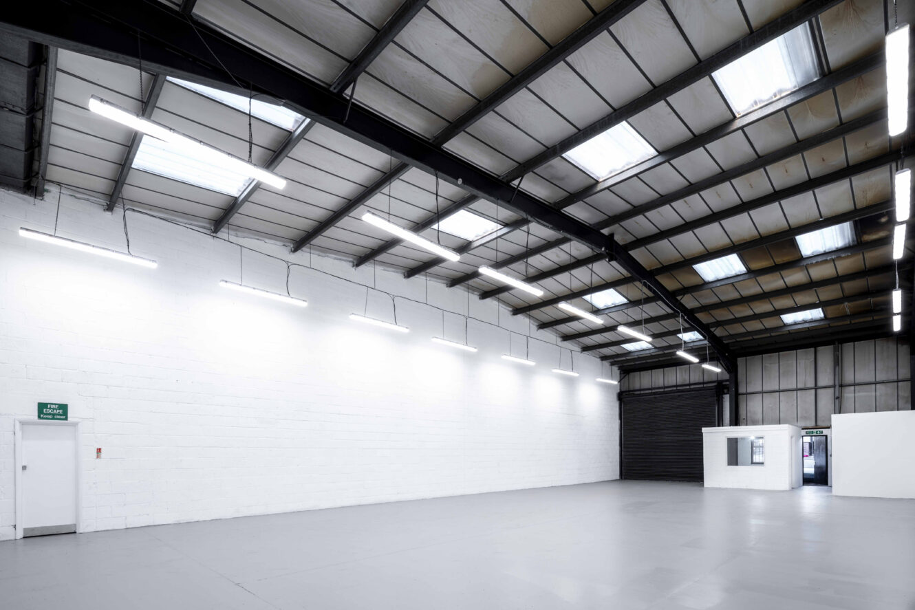 Large, empty warehouse with white walls, gray concrete floor, skylights, bright ceiling lights, a white door on the left, and a small white office room on the right.
