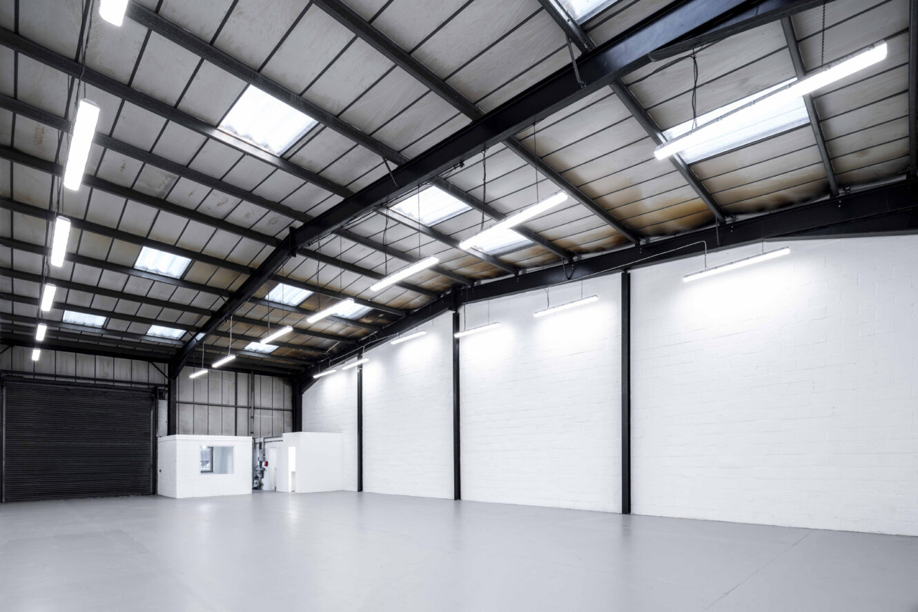 Large, empty industrial warehouse with high ceilings, white walls, concrete floor, overhead lights, and a small white office structure in the corner.