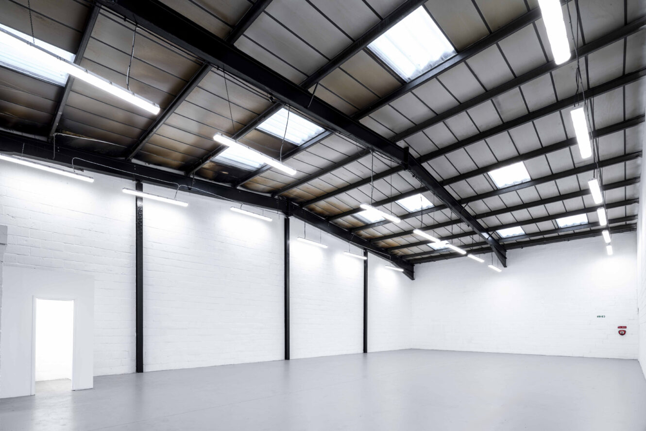 Interior of an empty industrial warehouse with white walls, a high ceiling with skylights, and fluorescent lighting.