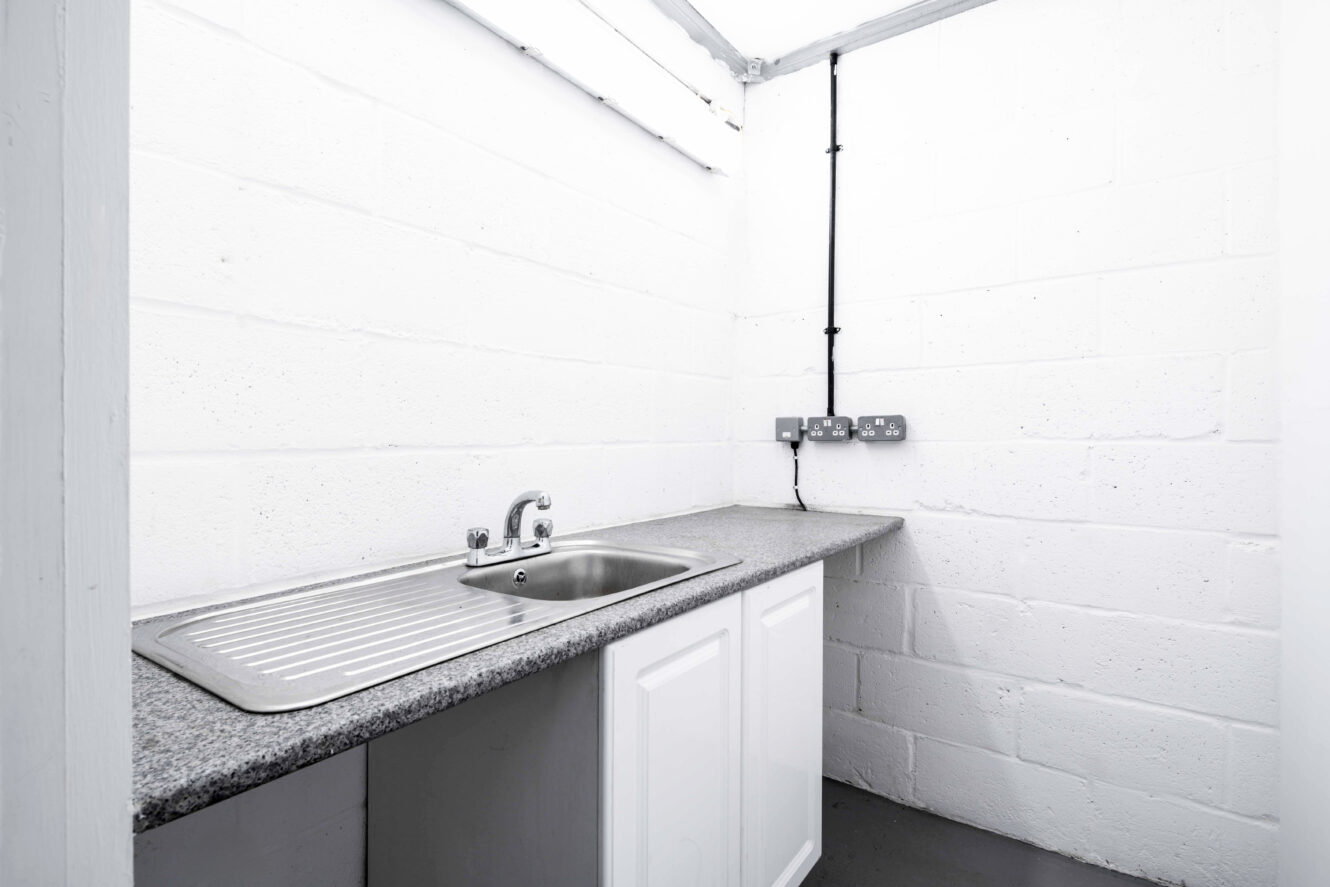 A small, minimalist kitchenette with a stainless steel sink, drainer, grey countertop, white cabinets, and exposed electrical outlets on white painted brick walls.