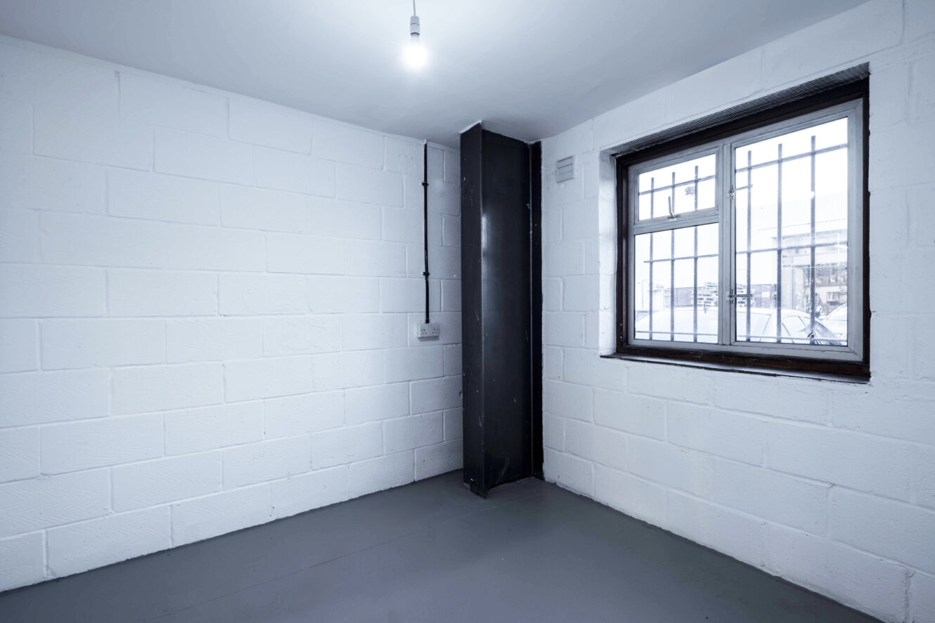 A small, empty room with white painted brick walls, a single barred window, a black vertical column, exposed wiring, and a grey floor under a ceiling light.