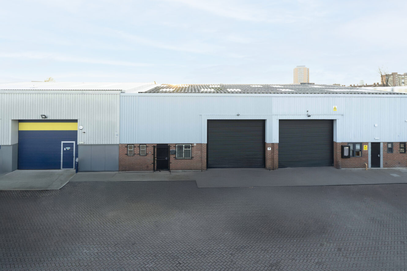 Industrial warehouse exterior with three large doors—two black rolling shutters and one blue—with a paved driveway in front.