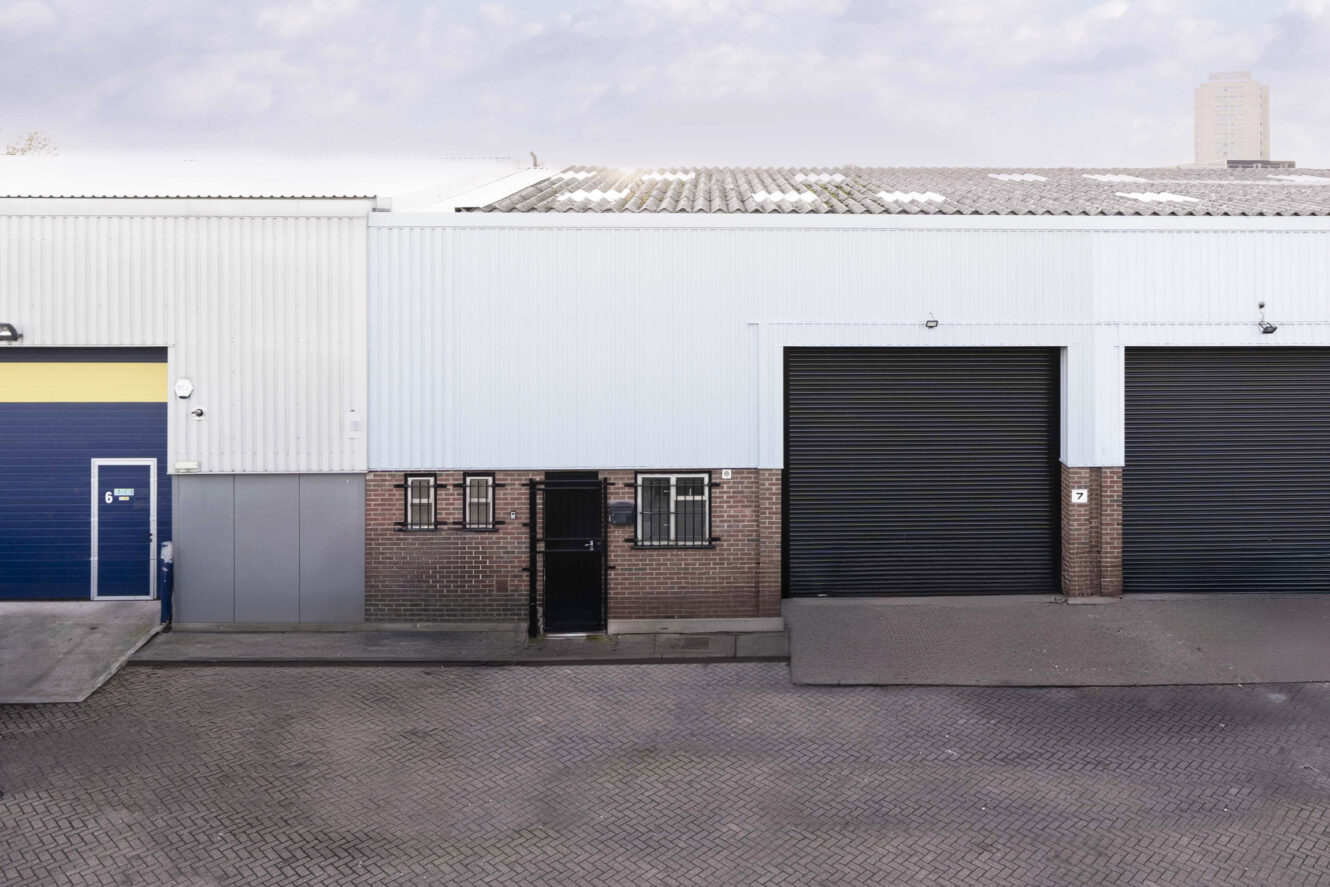 Industrial building with two large black roller doors, a blue entry door, small barred windows, and a brick-paved driveway in front.