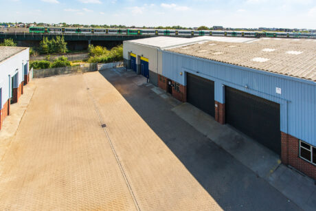 Industrial warehouse buildings with closed roller shutters and a paved yard; a train passes on elevated tracks in the background under a clear sky.