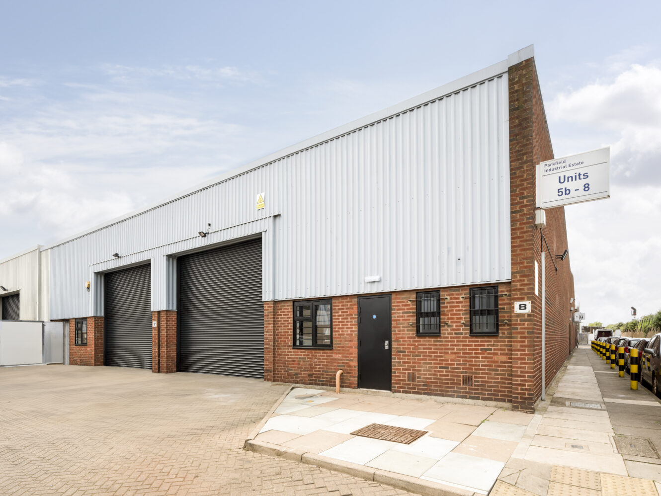 A modern industrial warehouse building with red brick and metal siding, featuring two large black roller doors, a single entry door, and unit signs visible.
