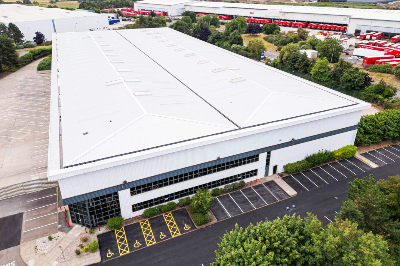 A large industrial warehouse with a white roof, surrounded by parking spaces, some marked for disabled access, and green landscaping. Other warehouses are visible in the background.