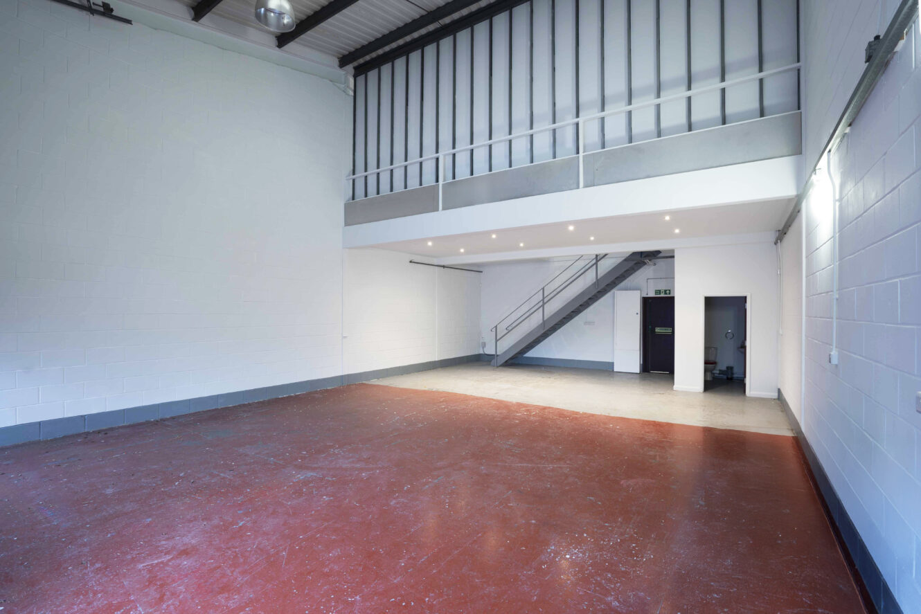 Empty industrial unit with red concrete floor, white walls, a mezzanine level, metal stairs, and fluorescent ceiling lights.