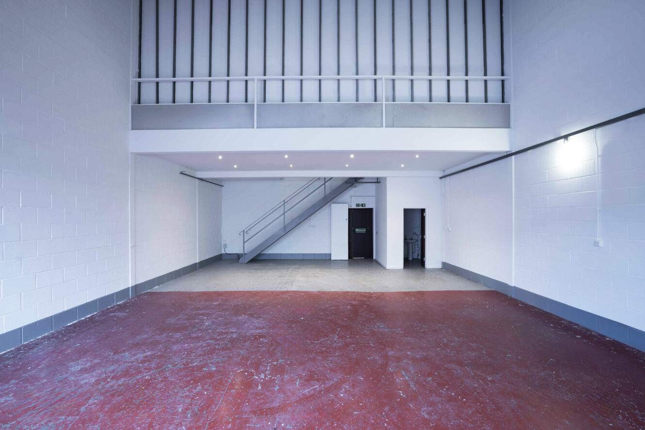 Empty industrial warehouse unit with red concrete floor, white brick walls, a mezzanine level, and a metal staircase leading to an upper floor.