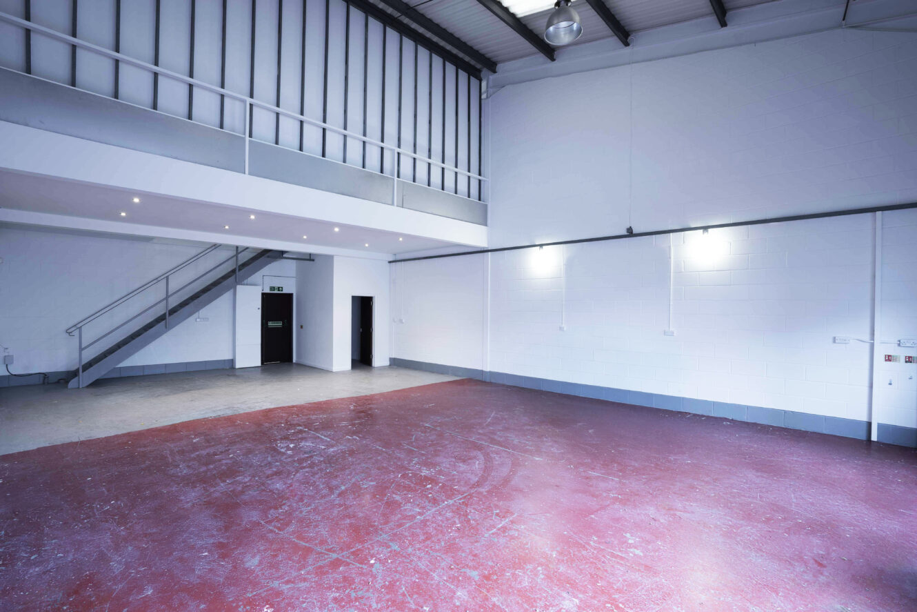 Empty industrial warehouse interior with red concrete floor, white walls, high ceiling, fluorescent lights, and a metal staircase leading to a mezzanine level.