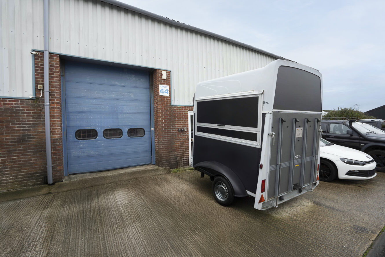 A black and white horse trailer is parked on a concrete surface outside a building with a blue roller shutter door and a white car nearby.