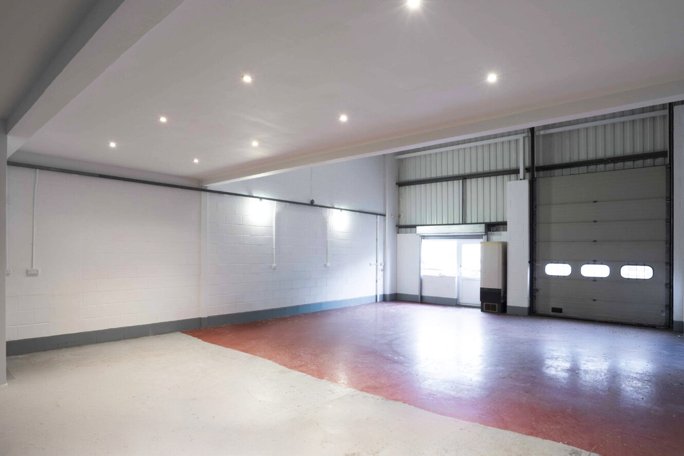 Empty industrial warehouse interior with white walls, red and white flooring, overhead lighting, and a large roller shutter door next to a window and small storage unit.