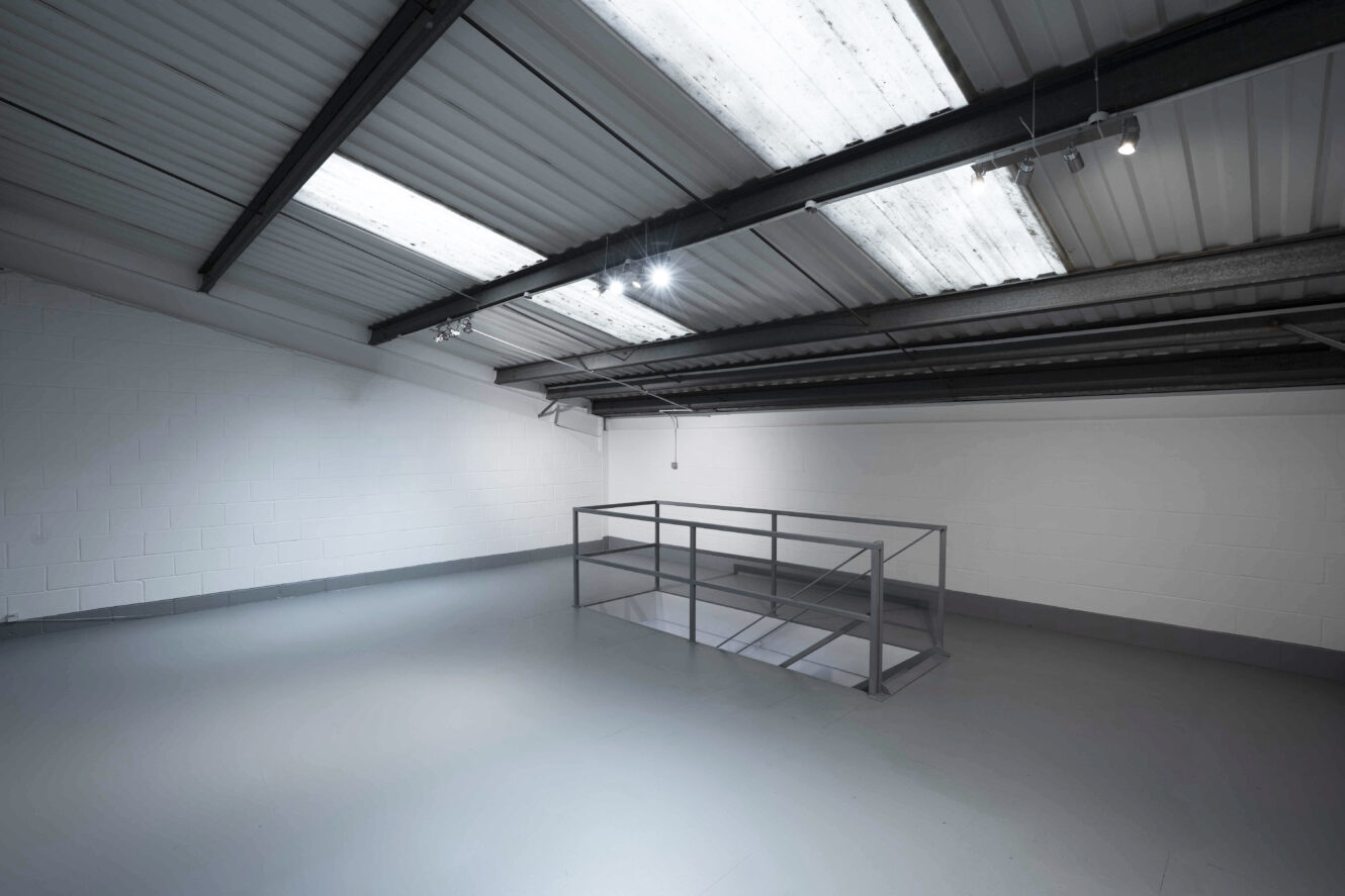 Empty industrial loft space with gray floors, white walls, and metal ceiling beams; a metal railing surrounds a staircase leading downstairs.
