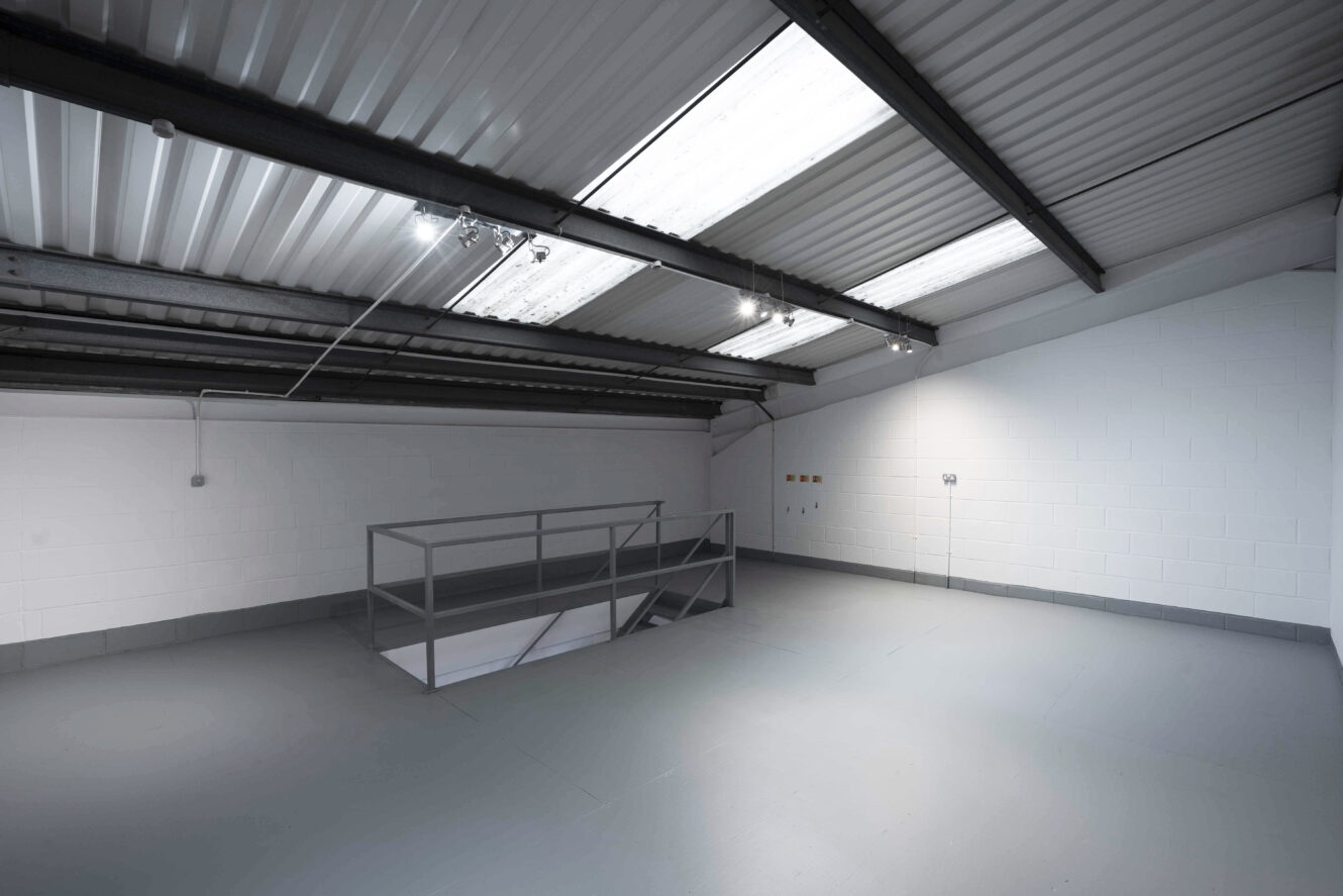 Empty industrial loft space with gray floor, white walls, metal railing around stairwell, and fluorescent lights on the corrugated ceiling.