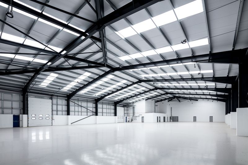 Interior of a large, empty warehouse with a high ceiling, metal beams, skylights, and several closed doors along the white walls.