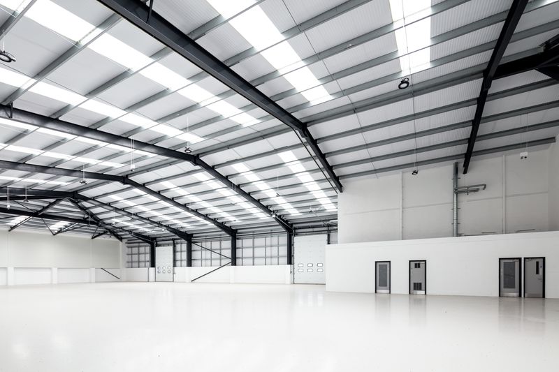 Spacious, empty industrial warehouse with high ceilings, white floors, and exposed steel beams, lit by natural and artificial lighting.