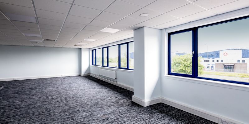Empty office space with gray carpet, white walls, and large windows overlooking an industrial area. Bright natural light fills the room.