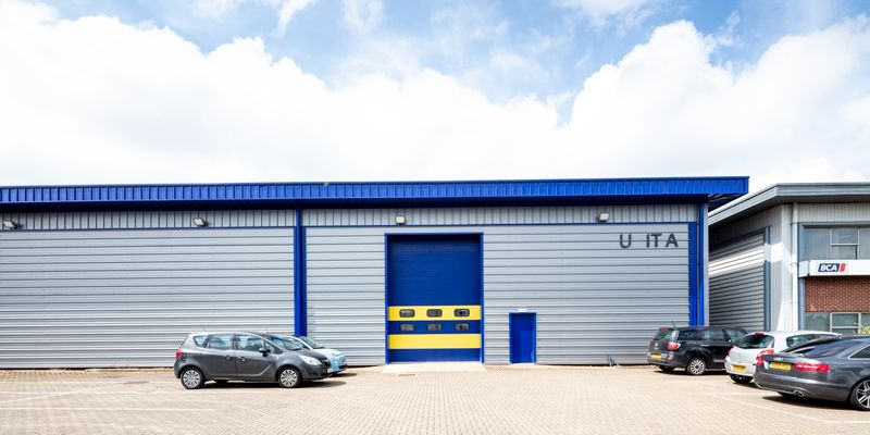 A modern industrial warehouse with blue and gray exterior, five parked cars, and a large blue-and-yellow loading door.