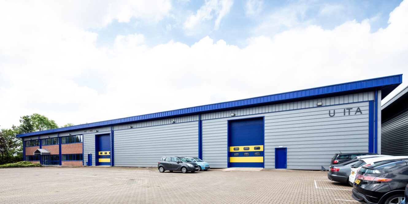 A large industrial warehouse with blue and silver exterior, yellow accents on doors, and several parked cars in front on a paved lot under a partly cloudy sky.