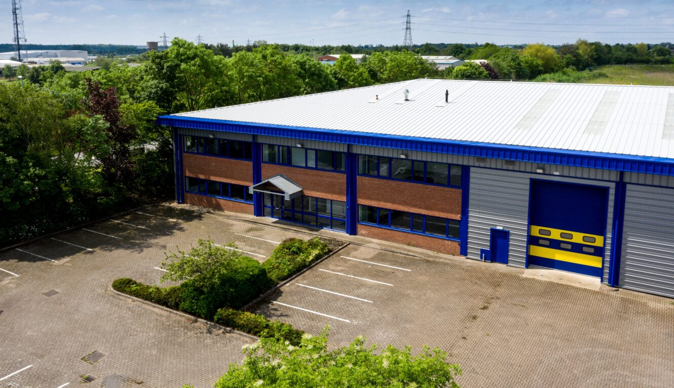 A large industrial building with blue accents, empty parking spaces, and surrounding greenery under a clear sky.