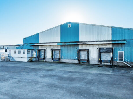 Large blue and white industrial warehouse with five loading dock doors, a small white office unit, and a wide concrete forecourt under a clear sky.