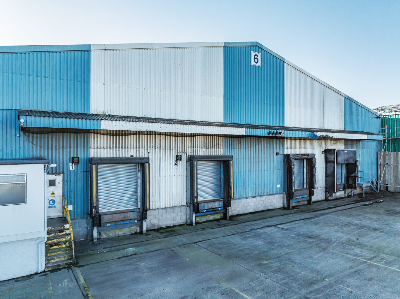 A large blue and white industrial warehouse with four closed loading dock doors and concrete steps on the left side, labeled with the number 6.