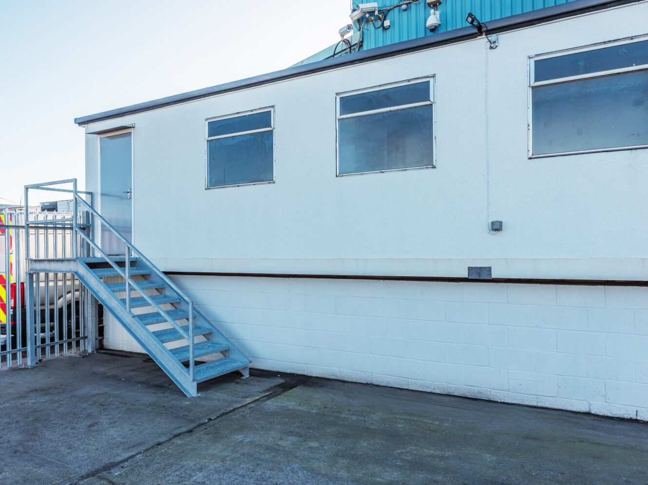 A white industrial building with three windows, a metal staircase leading to a door, and a concrete ground in front.