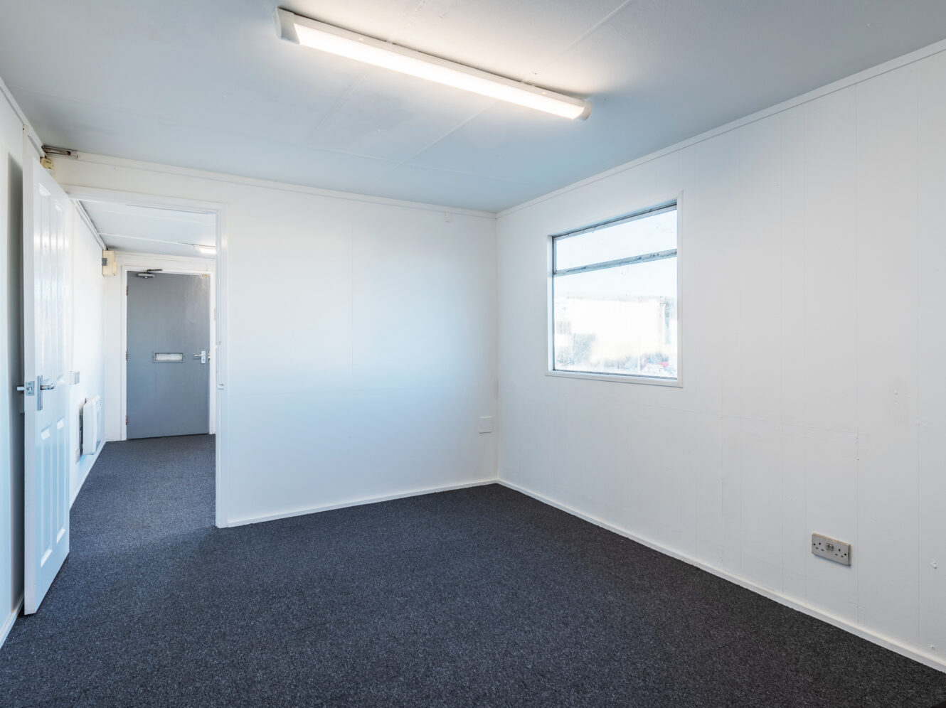 Empty room with white walls, a window, grey carpet, and fluorescent ceiling light; adjoining hallway and doors visible in background.