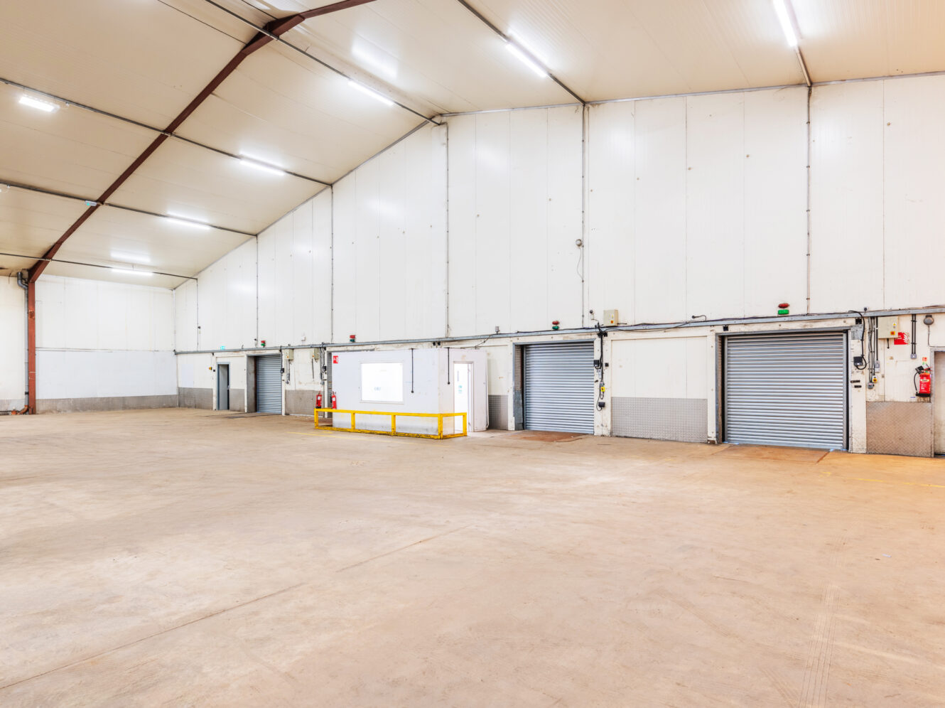 Interior of a large, empty warehouse with white walls, high ceiling, concrete floor, and three roller shutter doors.