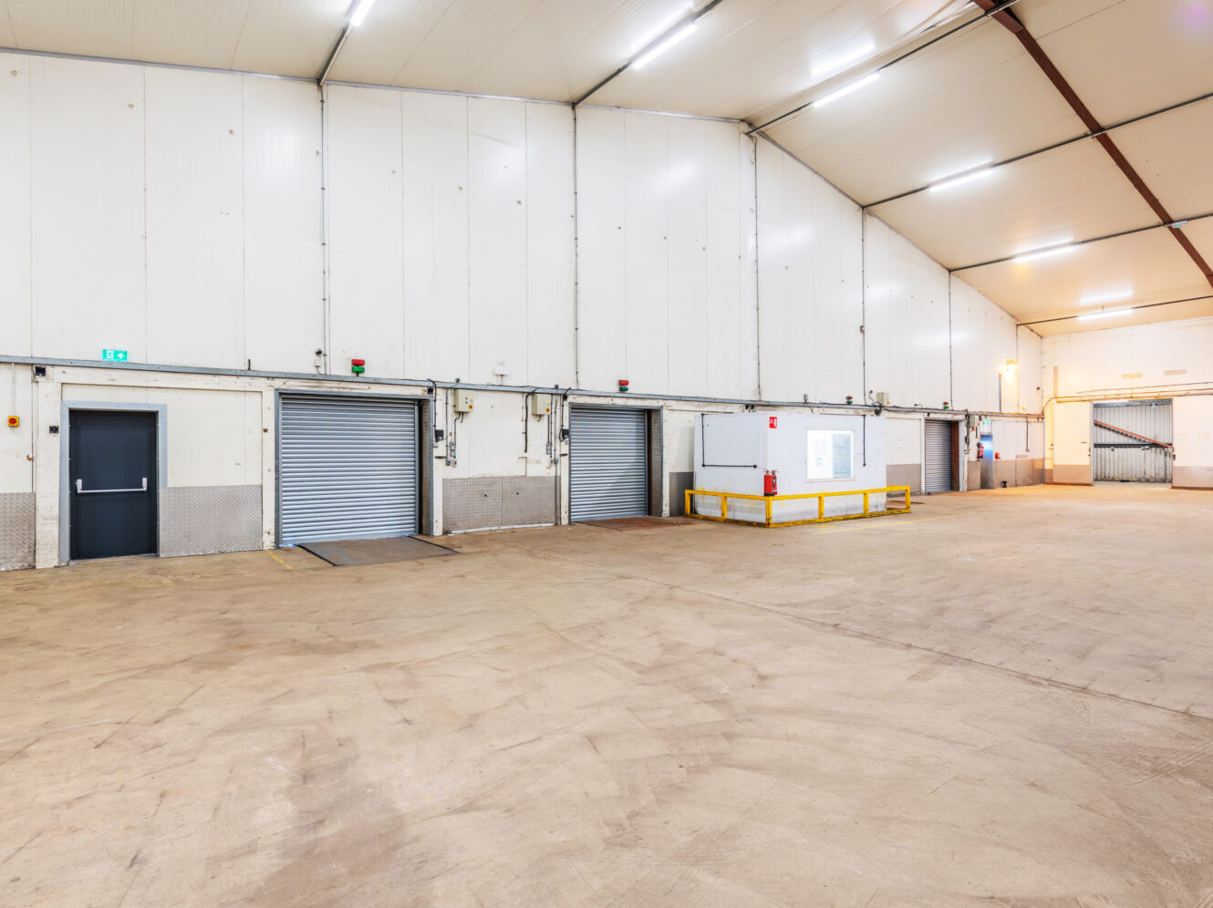Large, empty warehouse interior with white walls, high ceiling, three metal roller doors, a black door, yellow safety barriers, and fluorescent lighting.